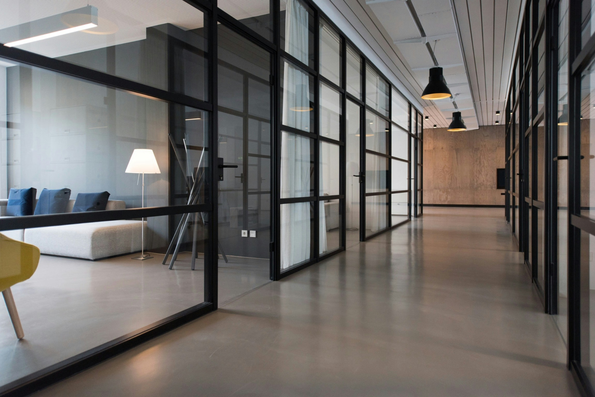 A quiet modern office corridor lined with glass-partitioned rooms and black pendant lights