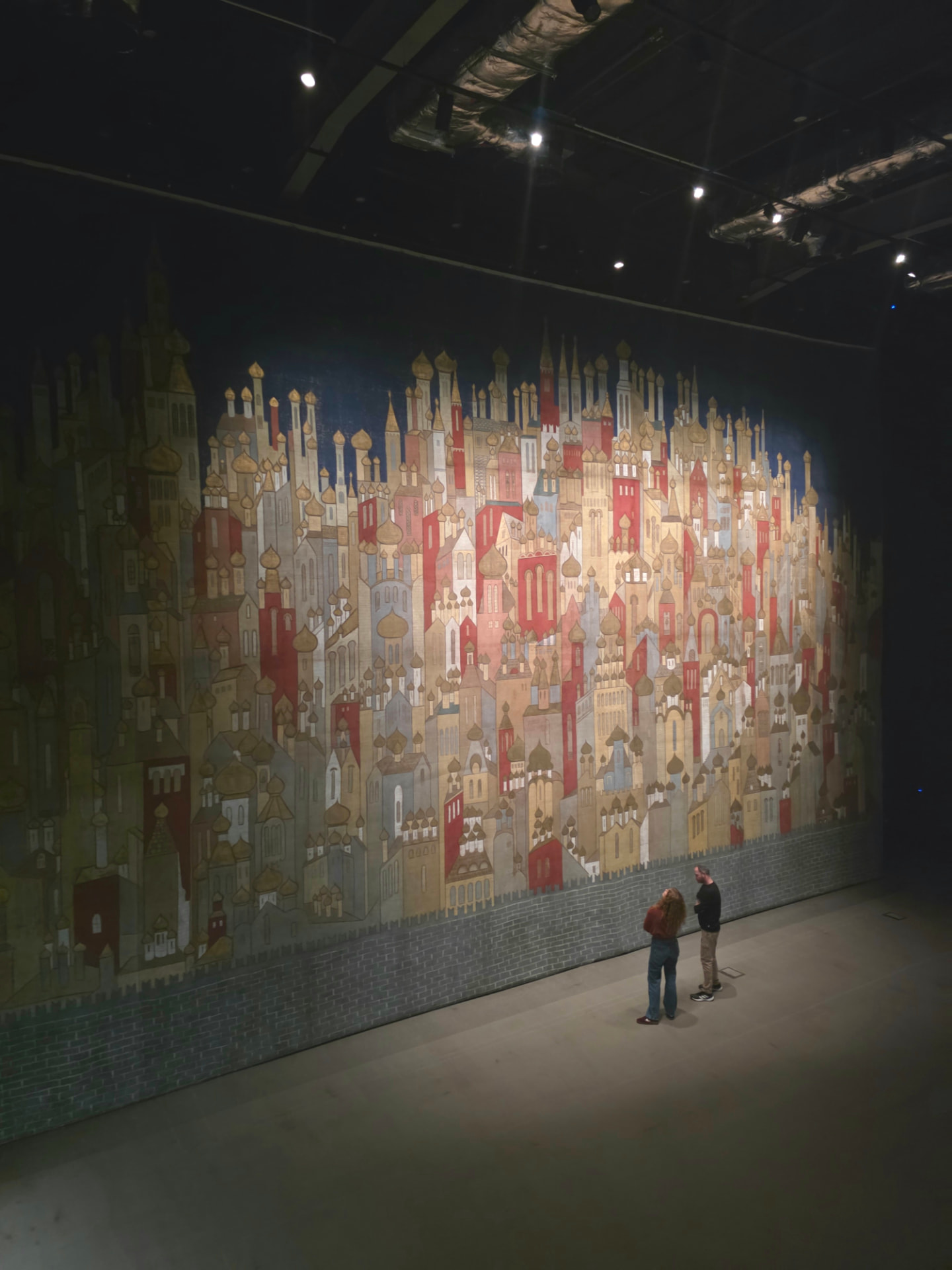 Two visitors standing before a towering mural of an intricate cityscape in a dimly lit gallery